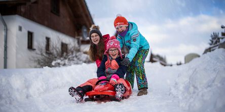 Ein Kind auf einem Schlitten und zwei Kinder dahinter schubsen es an.