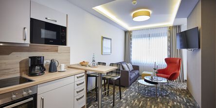 Offene Küche mit Wohn- und Essbereich mit rotem Sessel, grauer Couch auf gemustertem Teppichboden und Fernseher