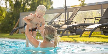 Frau im Pool hebt lachenden Jungen in die Luft, während Sonnenliegen im Hintergrund stehen