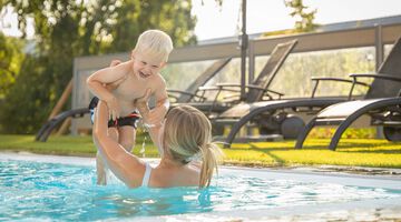 Frau im Pool hebt lachenden Jungen in die Luft, während Sonnenliegen im Hintergrund stehen