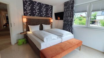 Schlafzimmer mit gemütlichem Doppelbett an einer Akzentwand mit Blumentapete neben einem Fenster mit gemusterten Vorhängen 