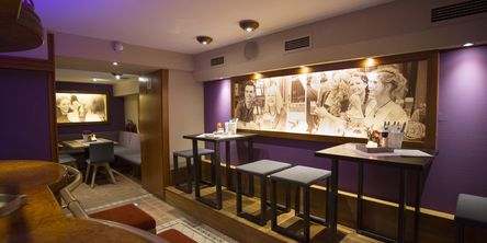 Einblick in die Bar mit lila gestrichenen Wänden, an denen große Bilder hängen, eingerichtet mit hohen Tischen und Hockern 