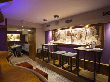 Einblick in die Bar mit lila gestrichenen Wänden, an denen große Bilder hängen, eingerichtet mit hohen Tischen und Hockern 