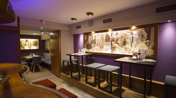Einblick in die Bar mit lila gestrichenen Wänden, an denen große Bilder hängen, eingerichtet mit hohen Tischen und Hockern 
