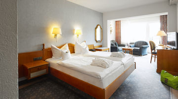 Großzügiges Doppelzimmer mit einem gemütlichem Bett neben einem Loungebereich mit zwei blauen Sesseln am Fenster