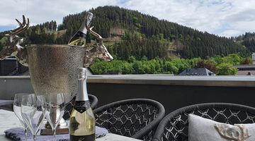 Silberner Sektkühler verziert mit zwei Ziegenbockköpfen auf einem Tisch vor grünen Bergen 