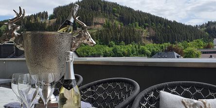 Silberner Sektkühler verziert mit zwei Ziegenbockköpfen auf einem Tisch vor grünen Bergen 