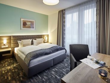 Gemütliches Doppelbett an einer türkisfarbenen Wand im Zimmer mit Schreibtisch und großem Fenster 