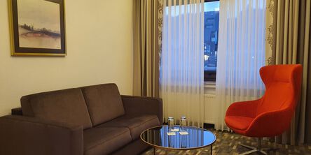 Dunkle Couch unter einem gerahmten Gemälde gegenüber eines roten Sessels an einem Glastisch mit zwei Trinkgläsern 