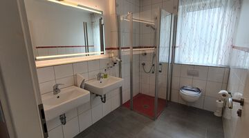 Weiß gefliestes Badezimmer mit zwei Waschbecken an einem beleuchteten Spiegel naben einer Dusche