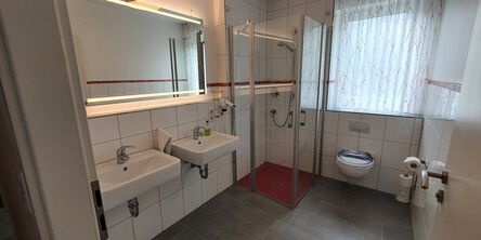 Weiß gefliestes Badezimmer mit zwei Waschbecken an einem beleuchteten Spiegel naben einer Dusche
