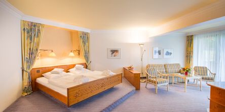Lichtdurchflutetes Zimmer mit einem großzügigem Bett auf einer Erhebung neben dem Wohnbereich mit karierter Couch zwischen zwei passenden Sesseln 