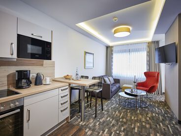 Offene Küche mit Wohn- und Essbereich mit rotem Sessel, grauer Couch auf gemustertem Teppichboden und Fernseher