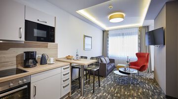 Offene Küche mit Wohn- und Essbereich mit rotem Sessel, grauer Couch auf gemustertem Teppichboden und Fernseher