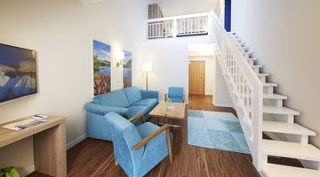 Der lichtdurchflutete Wohnraum mit blauen Sesseln und weißer Treppe im Galeriezimmer des Göbel Seehotels  Diemelsee