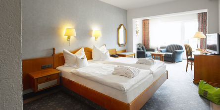 Großzügiges Doppelzimmer mit einem gemütlichem Bett neben einem Loungebereich mit zwei blauen Sesseln am Fenster