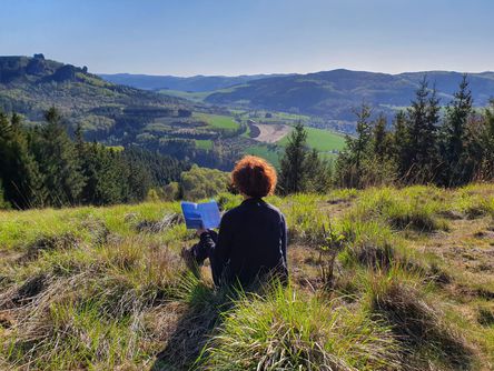 Frau sitzt auf einem Berggipfel mit Blick über die Landschaft und liest ein Buch 