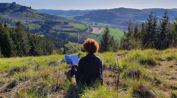 Frau sitzt auf einem Berggipfel mit Blick über die Landschaft und liest ein Buch 