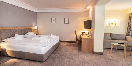 Großzügiges Zimmer mit einem großen Doppelbett, einem Schreibtisch unter einem Wandfernseher und einem abgetrennten Loungebereich mit grauer Couch