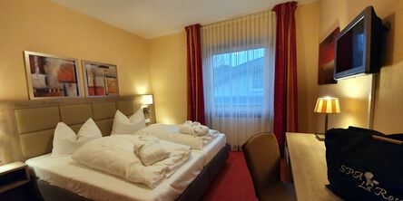Doppelzimmer mit gemütlichem Bett, gefalteten Bademänteln und zwei bunten Gemälden über dem Kopfteil neben den Fenster