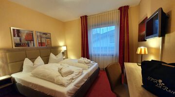 Doppelzimmer mit gemütlichem Bett, gefalteten Bademänteln und zwei bunten Gemälden über dem Kopfteil neben den Fenster